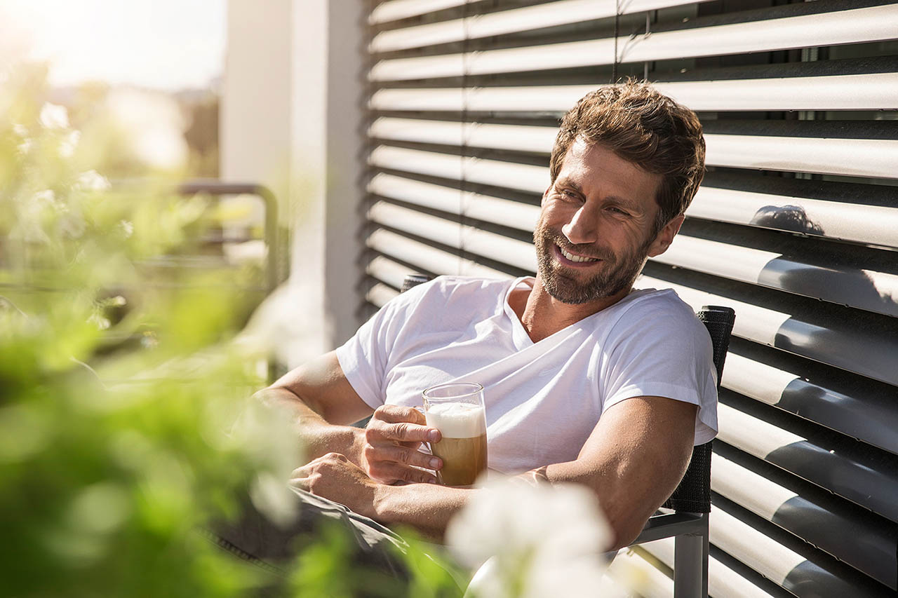 Männliches Model sitzt in einem Sessel auf der Terrasse mit einem Kaffee und lächelt